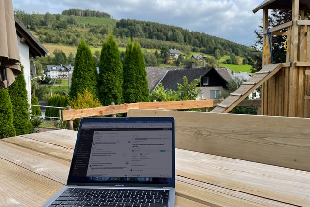 Ein geöffneter Laptop steht auf einem Gartentisch aus Holz. Im Hintergrund sind ein Spielplatz und Berge mit Wald und Wiesen zu sehen.